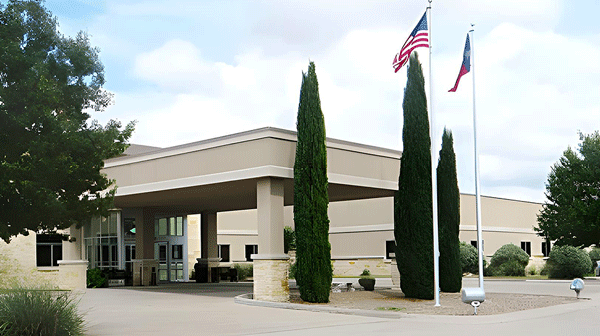 Iraan General Hospital - Texas Hospital Association Entrance to a beige commercial building with a porte-cochère, tall trees, and US and Texas flags on flagpoles outside.