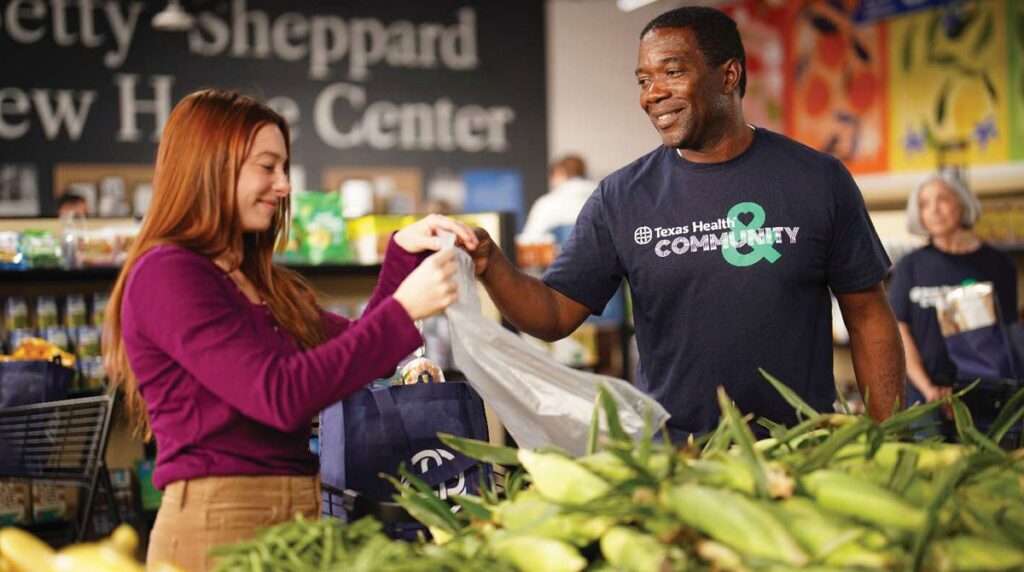 Texas Health Resources, 2026 Excellence In Community Service Award Winner - Texas Hospital Association Smiling man in a Texas Health Community shirt hands a bag to a woman at a grocery store donation event near a produce display.