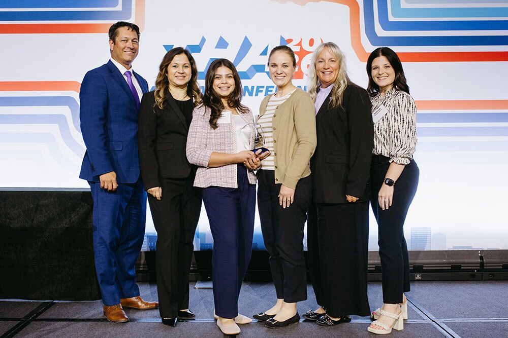 Bill Aston Award for Quality, Honorable Mention went to CHRISTUS Santa Rosa – Westover Hills, San Antonio - Texas Hospital Association Six professionals posing on a stage for an award photo, with one woman holding a glass trophy and a geometric backdrop behind them.