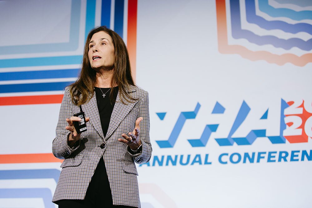 THA Board Chair Cris C. Daskevich, FACHE, CEO of CHRISTUS Children’s and SVP of CHRISTUS Health, spoke during the Awards Luncheon - Texas Hospital Association Female speaker on stage gesturing while presenting at a conference backdrop with bold geometric designs and the words'Annual Conference' visible.