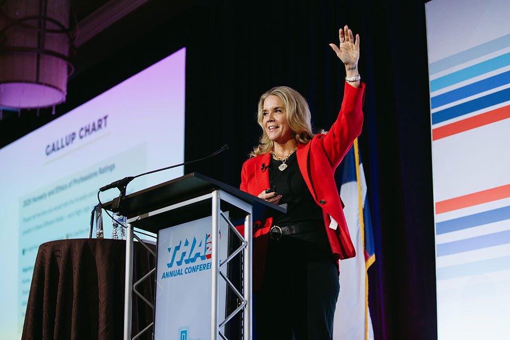 Jenifer Sarver, principal of Sarver Strategies, was the opening speaker the THA 2026 Annual Conference - Texas Hospital Association Woman in a red blazer speaks at a podium on stage, waving to the audience during a conference presentation (THA Annual Conference).