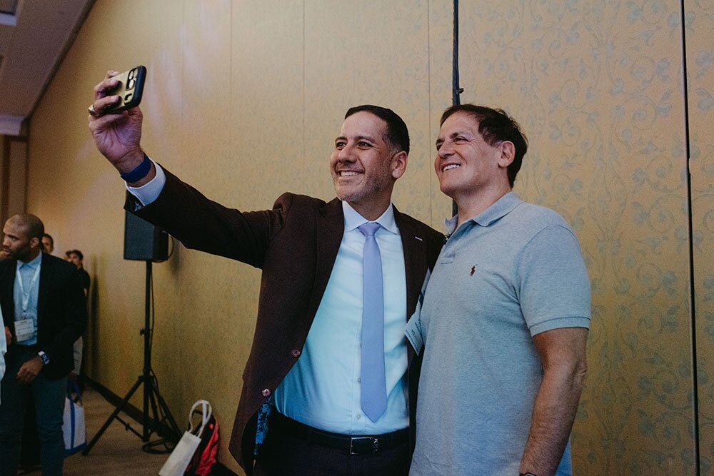 Laredo Medical Center CEO Jorge Leal took a selfie with Mark Cuban, co-founder of Cost Plus Drug Company - Texas Hospital Association Two men pose for a selfie at a conference, one in a suit and lavender tie and the other in a gray polo shirt, smiling together.
