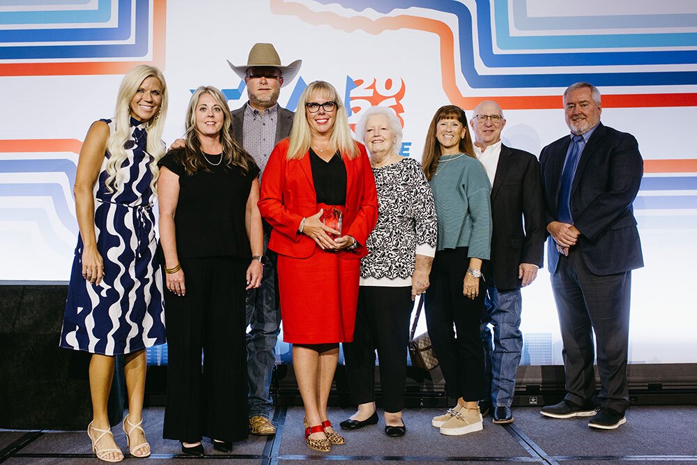 Pioneer Award winner Candice Smith (center) - Texas Hospital Association Eight adults posing on a stage at an event; a woman in a red suit holds a glass trophy against a colorful geometric backdrop.