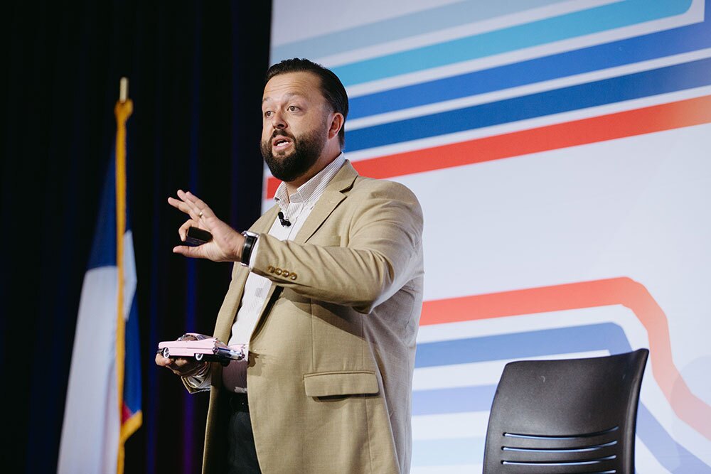 Ryan Campbell, founder of JoyAudit, closed the conference with “What’s Your Pink Cadillac?” - Texas Hospital Association Bearded man in a tan suit speaks on stage, holding a small car model and gesturing toward an audience, with a striped backdrop.