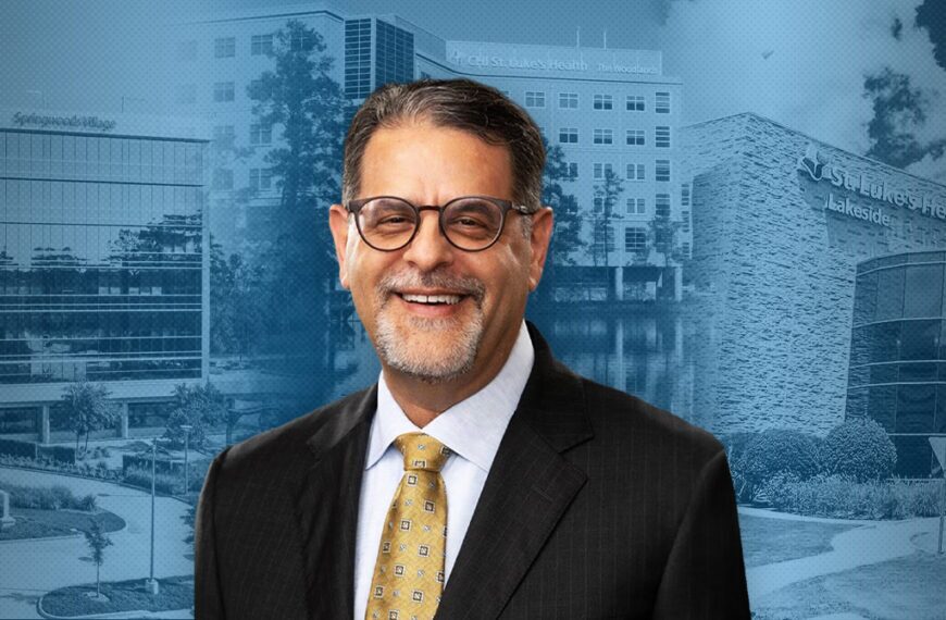 Smiling middle-aged man in a dark suit and yellow tie stands in front of a blue-tinted medical campus backdrop.
