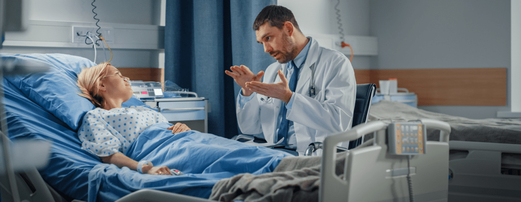 Patient Safety Organization - Texas Hospital Association Doctor in a white coat explains something to a patient lying in a hospital bed with medical equipment nearby.