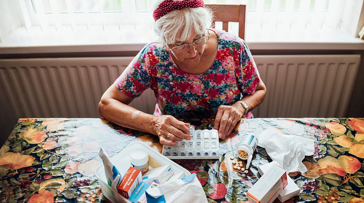 Elderly woman in a floral shirt sorting pills into a weekly pill organizer at a table full of medications and boxes.