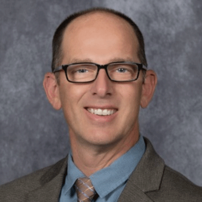Professional headshot of a smiling man wearing glasses, a blue shirt, brown blazer, and plaid tie against a gray backdrop.