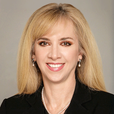 Head-and-shoulders portrait of a smiling blonde woman wearing a black blazer and pearl necklace.