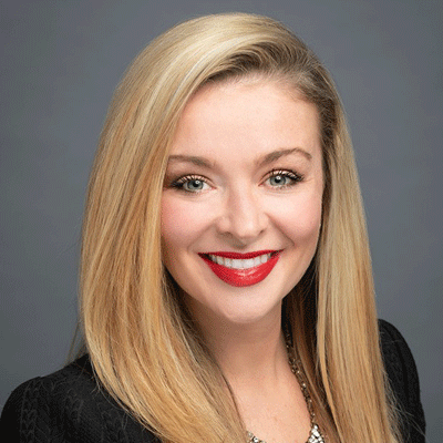 Professional headshot of a smiling blonde woman in a black blazer against a gray background.