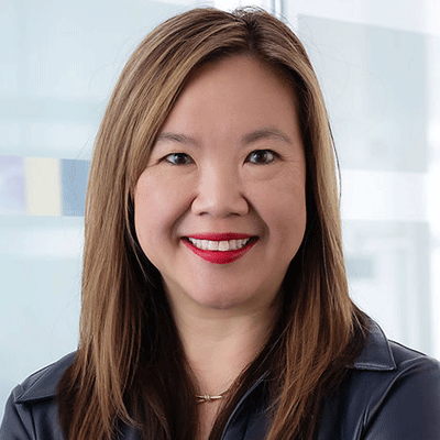 Portrait of a smiling woman with shoulder-length brown hair, wearing a dark blue blouse, in a bright office setting.
