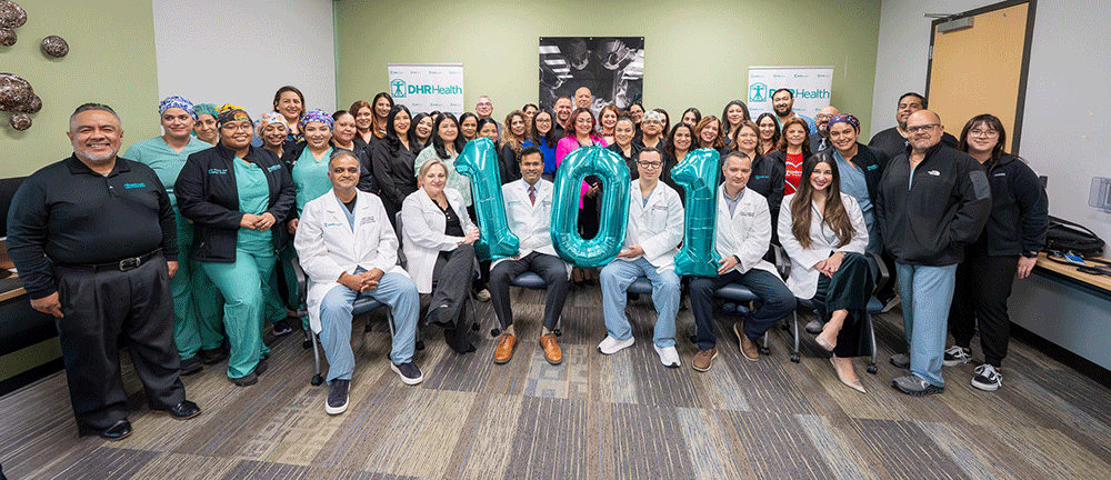 Group Photo of DHR Health Kidney Transplant Team - Texas Hospital Association Group of doctors and staff posing in a medical office, holding teal'100' balloons to celebrate a milestone, with DHR Health banners in the background.