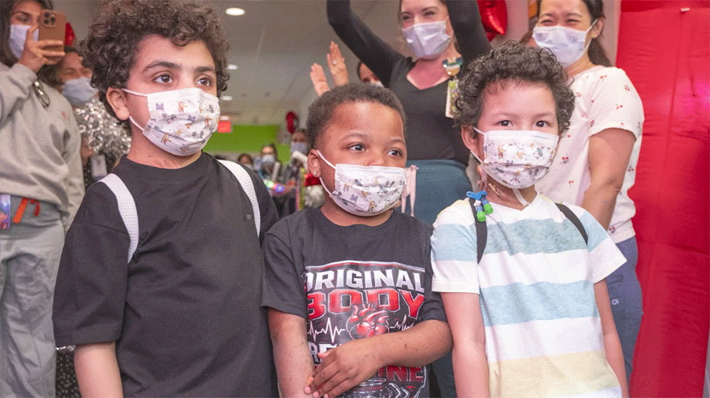 Three young boys who received heart transplants at Texas Children's Hospital - Texas Hospital Association Three children wearing patterned face masks stand together at an indoor event, smiling despite the masks, with cheering adults in the background.