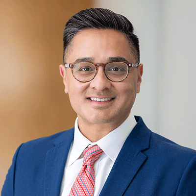 Portrait of a smiling man in a blue suit, white shirt, pink tie and glasses, in an office setting.