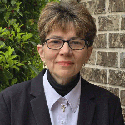Portrait of a woman with short light brown hair and glasses, wearing a dark blazer and white shirt with a cross necklace, standing beside a brick wall.