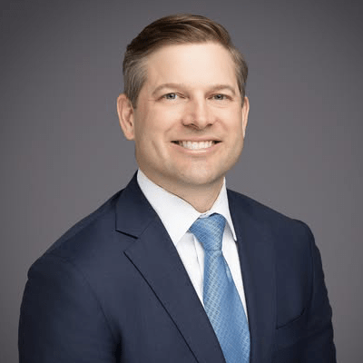 Professional headshot of a smiling man in a navy suit and light blue tie against a gray background.