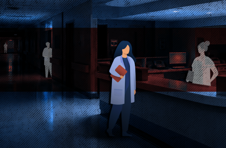 Healthcare worker in a blue coat holding a folder, standing in a dim hospital hallway.