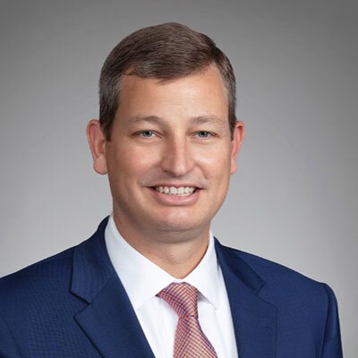 Professional headshot: smiling man in a navy suit with an orange tie against a gray backdrop.
