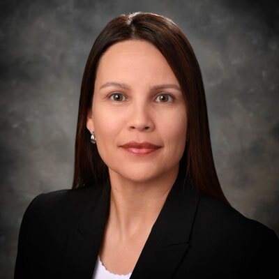 Professional headshot of a woman with dark brown hair in a black blazer, against a gray studio background.