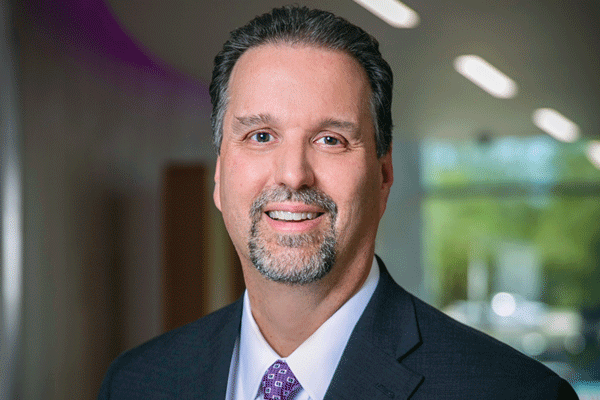 Chris Siebenaler - Texas Hospital Association Professional headshot of a smiling man in a dark suit and purple tie in an office hallway.