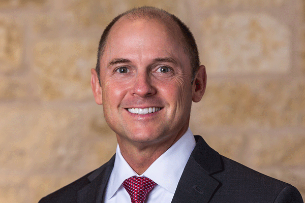 Cory Edmondson - Texas Hospital Association Smiling man in a dark suit and red tie, posed against a beige stone wall (professional headshot).