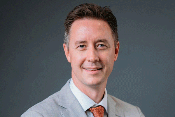 Daniel Miller - Texas Hospital Association Head-and-shoulders portrait of a smiling man in a light gray suit and orange tie against a neutral gray background