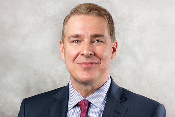 Donald Baker - Texas Hospital Association Head-and-shoulders portrait of a smiling man in a navy suit, checkered shirt, and red tie against a gray background
