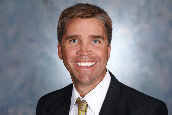 Eric Hamon - Texas Hospital Association Professional headshot of a smiling man in a black suit and yellow patterned tie against a neutral blue-gray backdrop.
