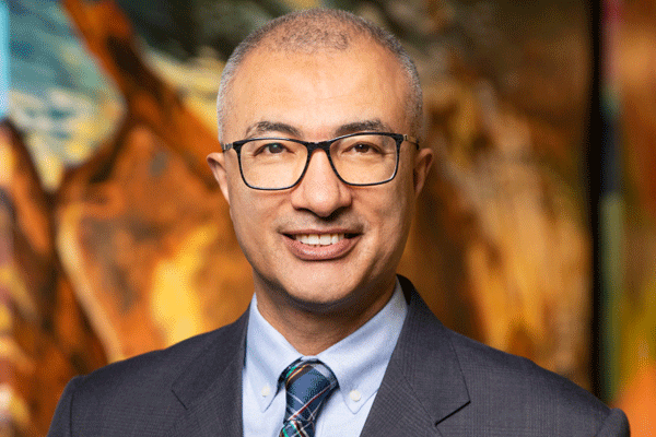 Esmaeil Porsa - Texas Hospital Association Professional head-and-shoulders portrait of a smiling man in a dark suit and glasses, with a blurred colorful background.
