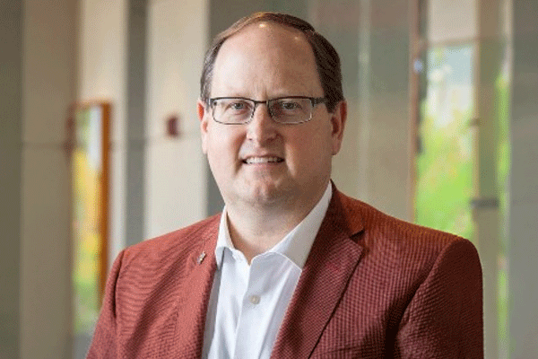 Gregory Haralson - Texas Hospital Association Professional headshot of a man with glasses wearing a white shirt and red blazer in a light office hallway.
