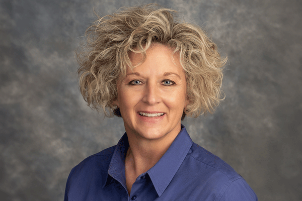 Holly Holcomb - Texas Hospital Association Smiling woman with short curly gray hair wearing a blue collared shirt, neutral studio backdrop behind her.