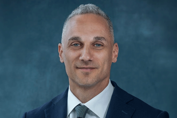 Jamie Youssef - Texas Hospital Association Professional headshot of a middle-aged man with short gray hair in a navy suit and teal tie against a blue backdrop.