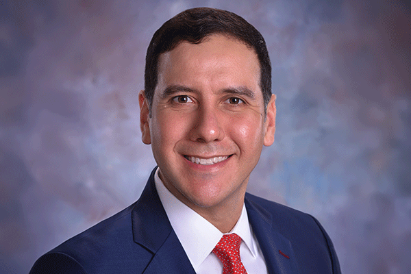 Jorge Leal - Texas Hospital Association Professional headshot of a smiling man in a navy suit, white shirt, red tie, against a blue-gray backdrop.