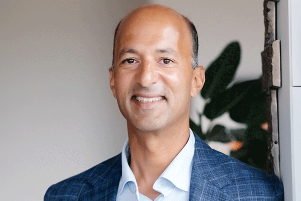 Sam Bagchi - Texas Hospital Association Professional portrait of a man in a blue blazer and white shirt smiling at the camera, indoors with plants in the background