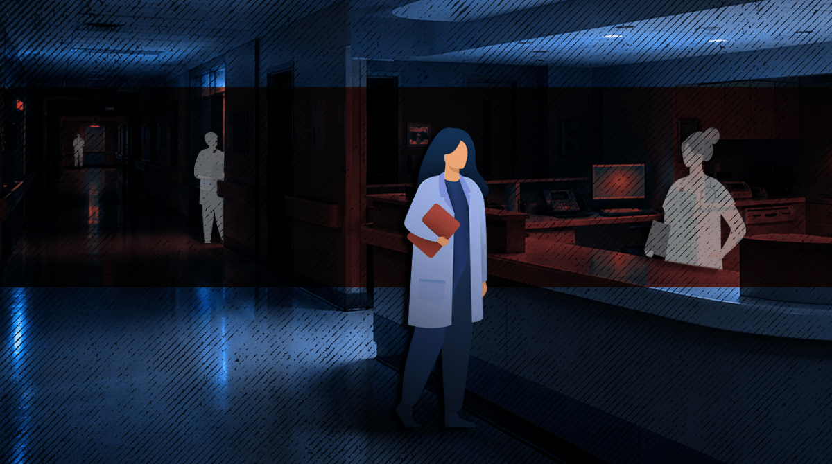 Healthcare worker in a blue coat holding a folder, standing in a dim hospital hallway.
