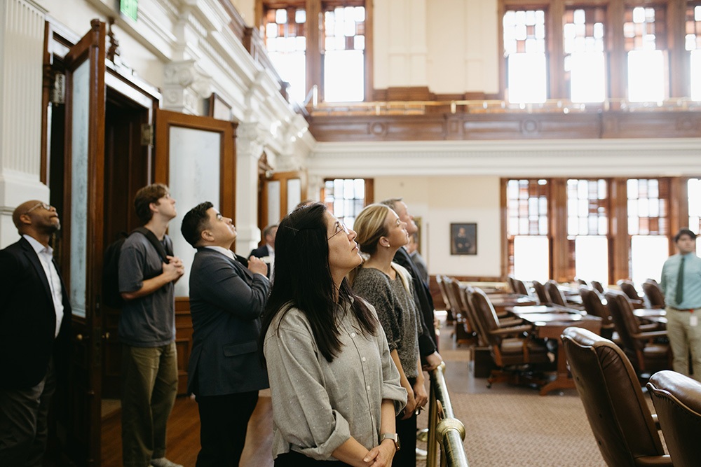 THA Fellows Class of 2026 visit the Texas Capitol - Texas Hospital Association Diverse group of professionals standing and looking up at the ornate ceiling in a wood-paneled legislative chamber in a historic meeting hall.