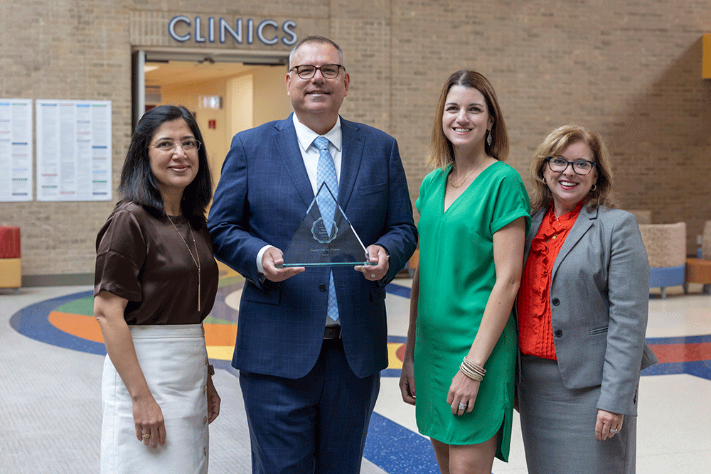 Angie Buckmeier, right - Texas Hospital Association Four professionals stand in a clinic lobby; a man in a blue suit holds a glass award and smiles, flanked by two women and a companion in business attire.