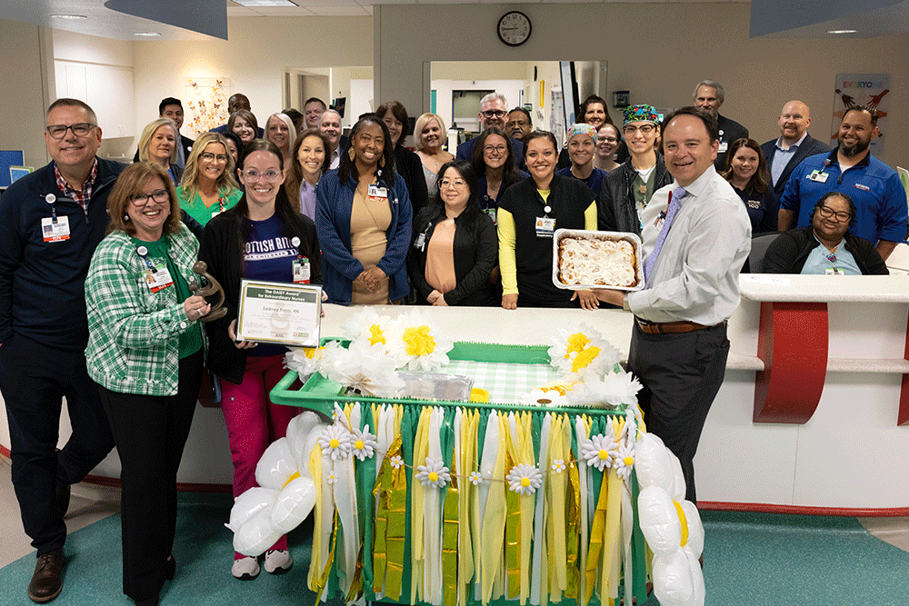 Angie Buckmeier and hospital staff celebrating a Daisy award - Texas Hospital Association Group of healthcare workers posing in a hospital lobby with a decorated table and cake for a celebration, one person holding a certificate and another a tray of baked goods.