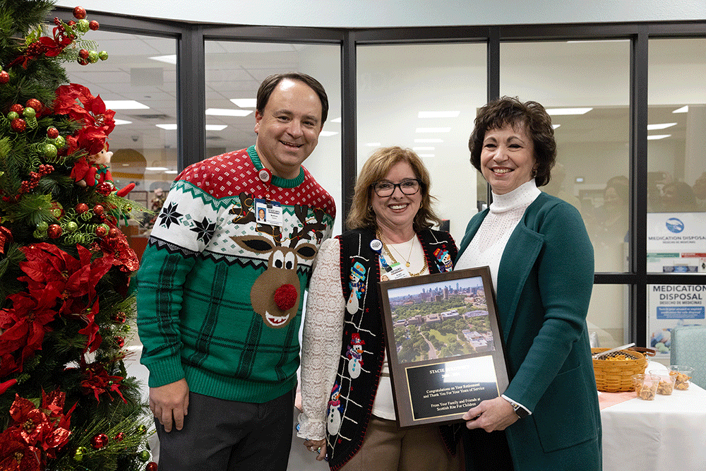 Angie Buckmeier - Texas Hospital Association Three adults smile for a photo beside a Christmas tree; the woman in the teal coat holds a framed award with another woman and a man wearing a festive sweater nearby.