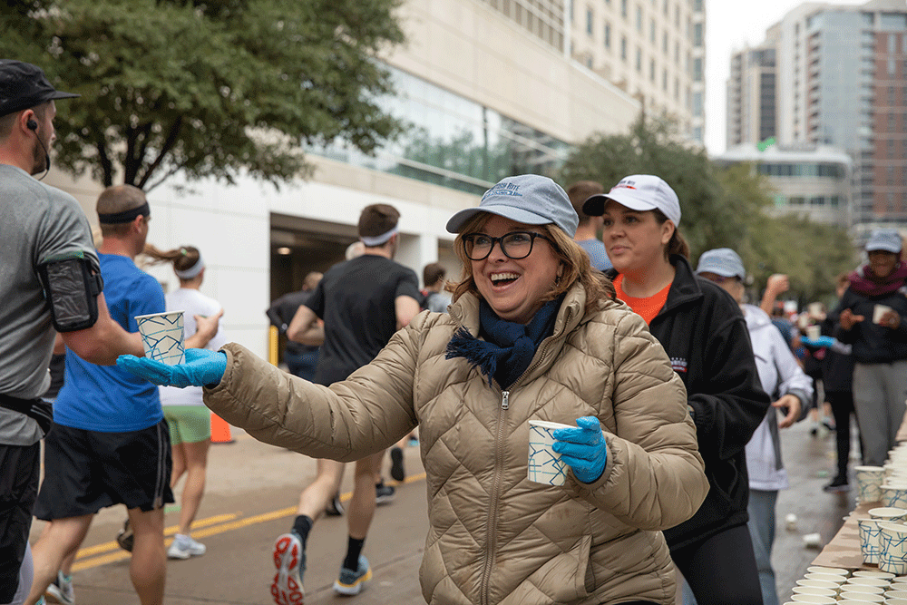 Angie Buckmeier handing out drinks to runners during the 2022 Dallas marathon - Texas Hospital Association Smiling woman in a beige jacket and cap hands a cup to a runner at an urban race aid station. Several volunteers and runners in the background.