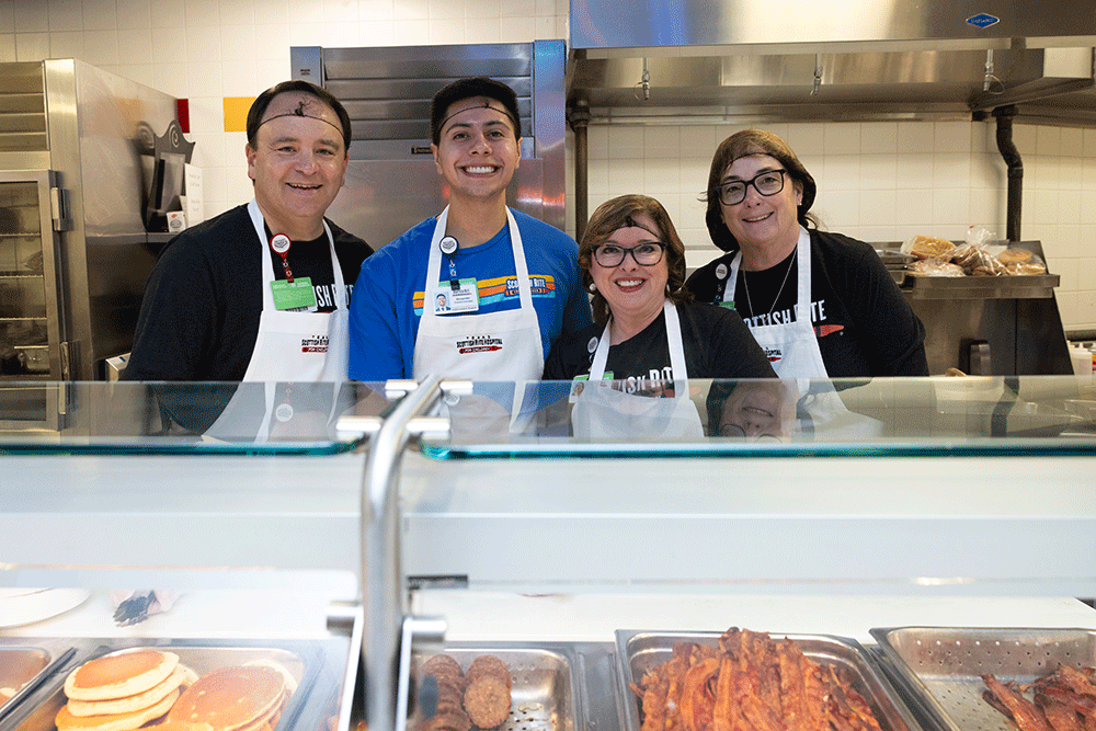 Angie Buckmeier, center right, participating in a pancake breakfast - Texas Hospital Association Four smiling coworkers behind a food counter wearing aprons in a busy kitchen setting.