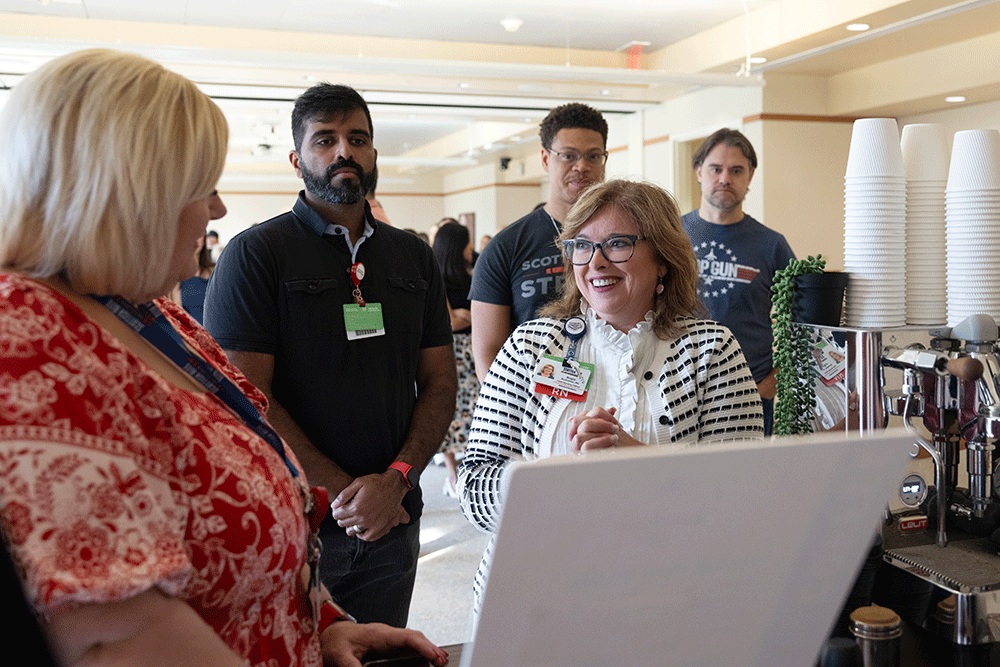 Angie Buckmeier during a Spirit Week Coffee Break - Texas Hospital Association Smiling woman barista with a name badge talks with a group of visitors at a coffee station; cups and espresso machine visible in the background.