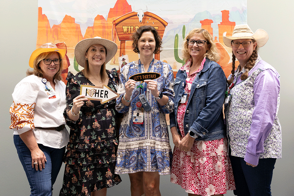 Angie Buckmeier, left, poses during Western Week - Texas Hospital Association Five women in cowboy hats and colorful outfits pose together at a western-themed event, one holding a sign that says'HER' and another 'LET'S PARTY' at center.