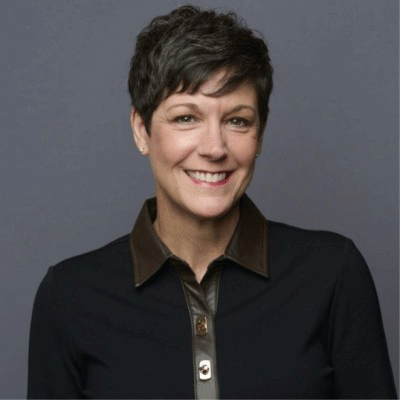 Denise Sutler - Texas Hospital Association Portrait of a smiling woman with short dark hair, wearing a black blazer over a brown collared shirt against a gray background.