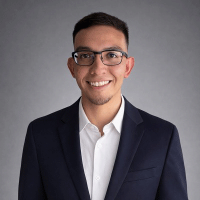 Spencer Estrella - Texas Hospital Association Professional headshot of a smiling man wearing a navy blazer and white shirt, with glasses, against a gray backdrop.
