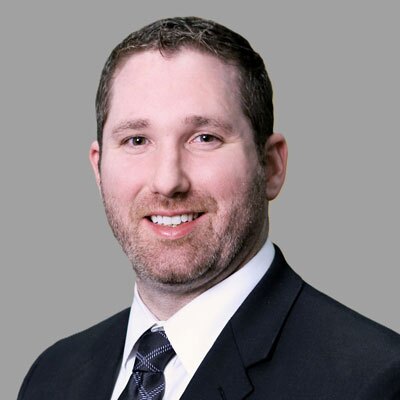 Andrew-Wilson - Texas Hospital Association Professional headshot of a smiling man in a dark suit and tie against a gray background.