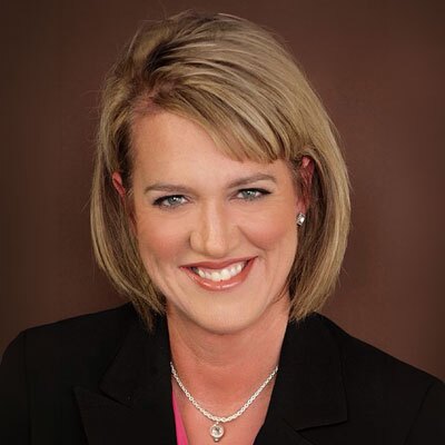 Wendi De Leon - Texas Hospital Association Professional headshot of a smiling blonde woman in a black blazer and pink blouse against a brown backdrop, wearing a silver necklace.