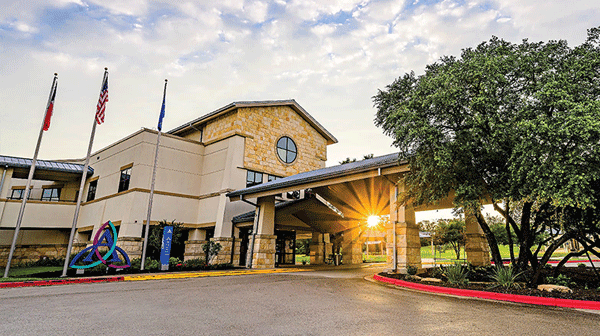Ascension Seton Southwest - Texas Hospital Association Hospital entrance with a covered driveway, flagpoles (US and state flag), stone facade, and a large tree on the right; sun glare near the doorway.