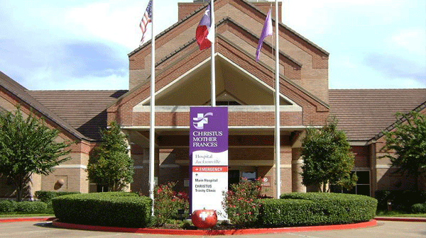Christus Mother Frances Hospital - Jacksonville - Texas Hospital Association Hospital entrance with three flagpoles and a CHRISTUS Mother Frances banner, brick building, and landscaped shrubs in front.