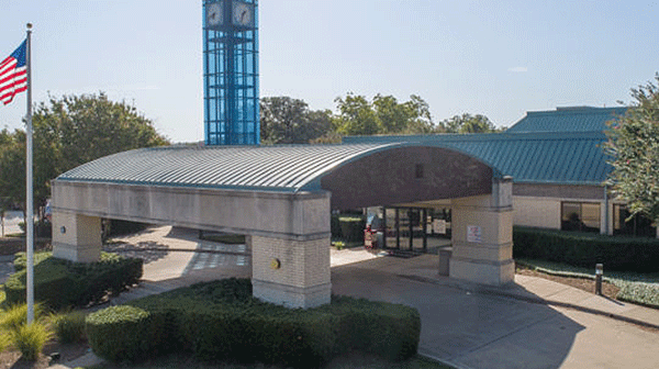 Glen Rose Medical Center - Texas Hospital Association Exterior view of a public building with a curved metal roof entrance, stone columns, a tall blue clock tower, and an American flag.